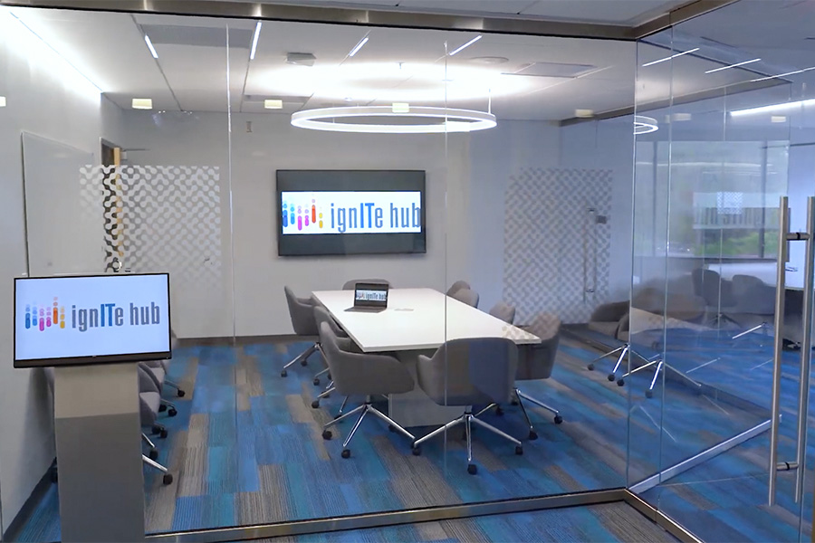 The ignite hub conference room is well lit with a white conference table in the middle surrounded by seats. There is a TV on the wall displaying the ignite hub logo